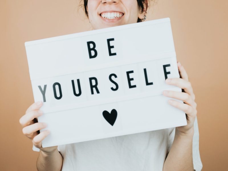 Young woman smiling to camera while holding a sign ( be yourself ) with positive vibes to camera. Cheering people concept.help and self help concept, mental health. color background with copy space.